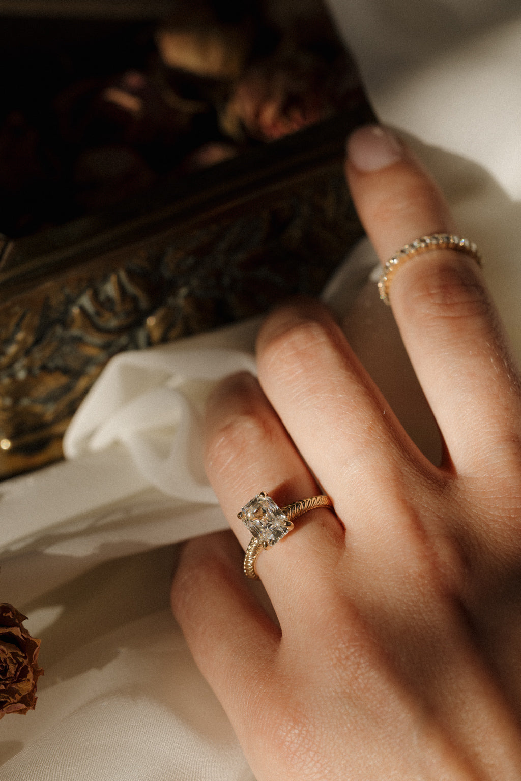 On-hand view of a yellow gold engagement ring with an antique emerald cut diamond in four claw prongs on a hand-engraved band, with a diamond band on the index finger, the hand resting on white fabric beside an antique-style brass jewelry box and a dried rose.