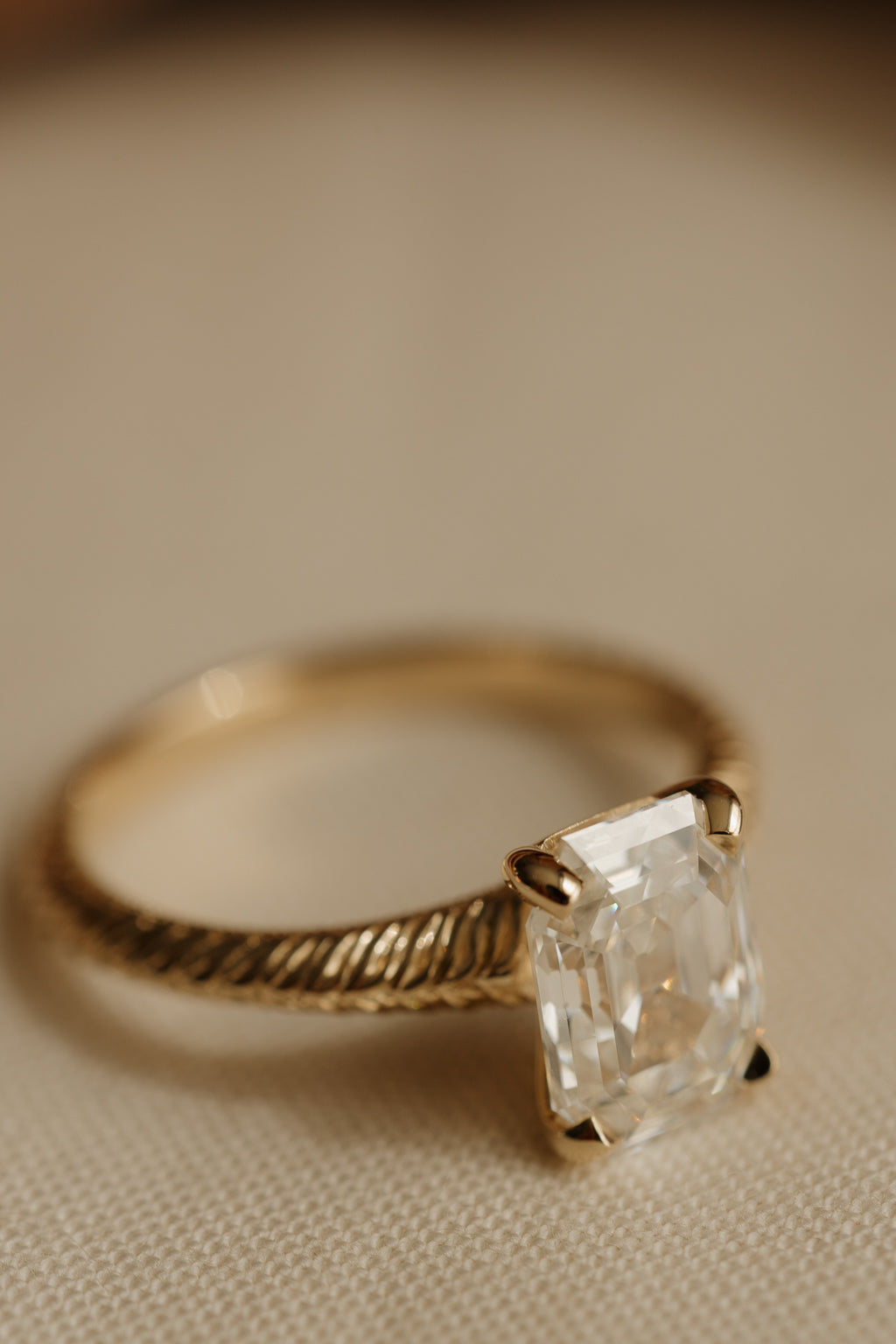Angled view of a yellow gold engagement ring with an antique emerald cut diamond in four claw prongs on a hand-engraved band, on cream woven linen.