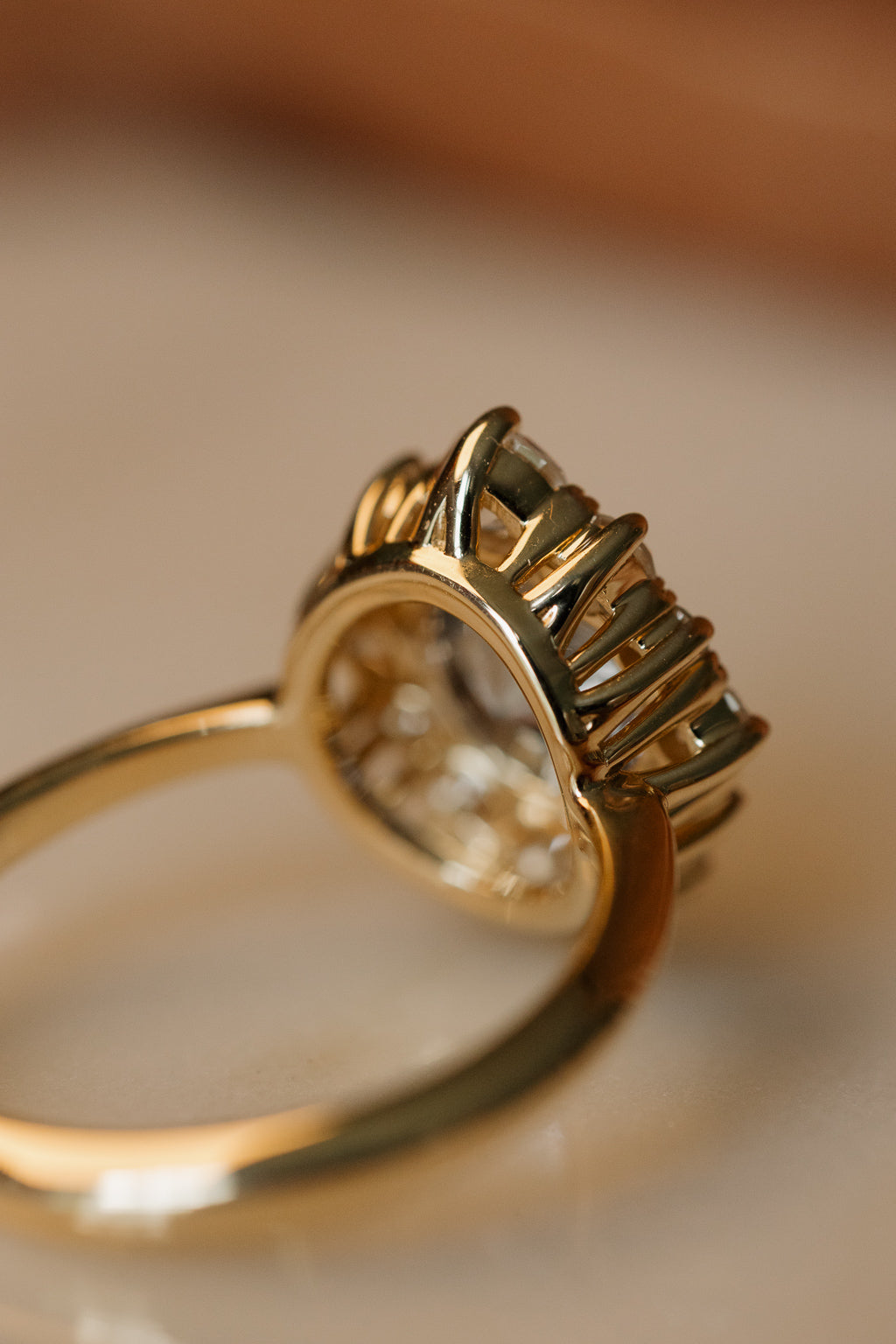 Side view of a yellow gold halo engagement ring from behind, showing the crown of claw prongs and open gallery beneath the stones, in warm, soft light.