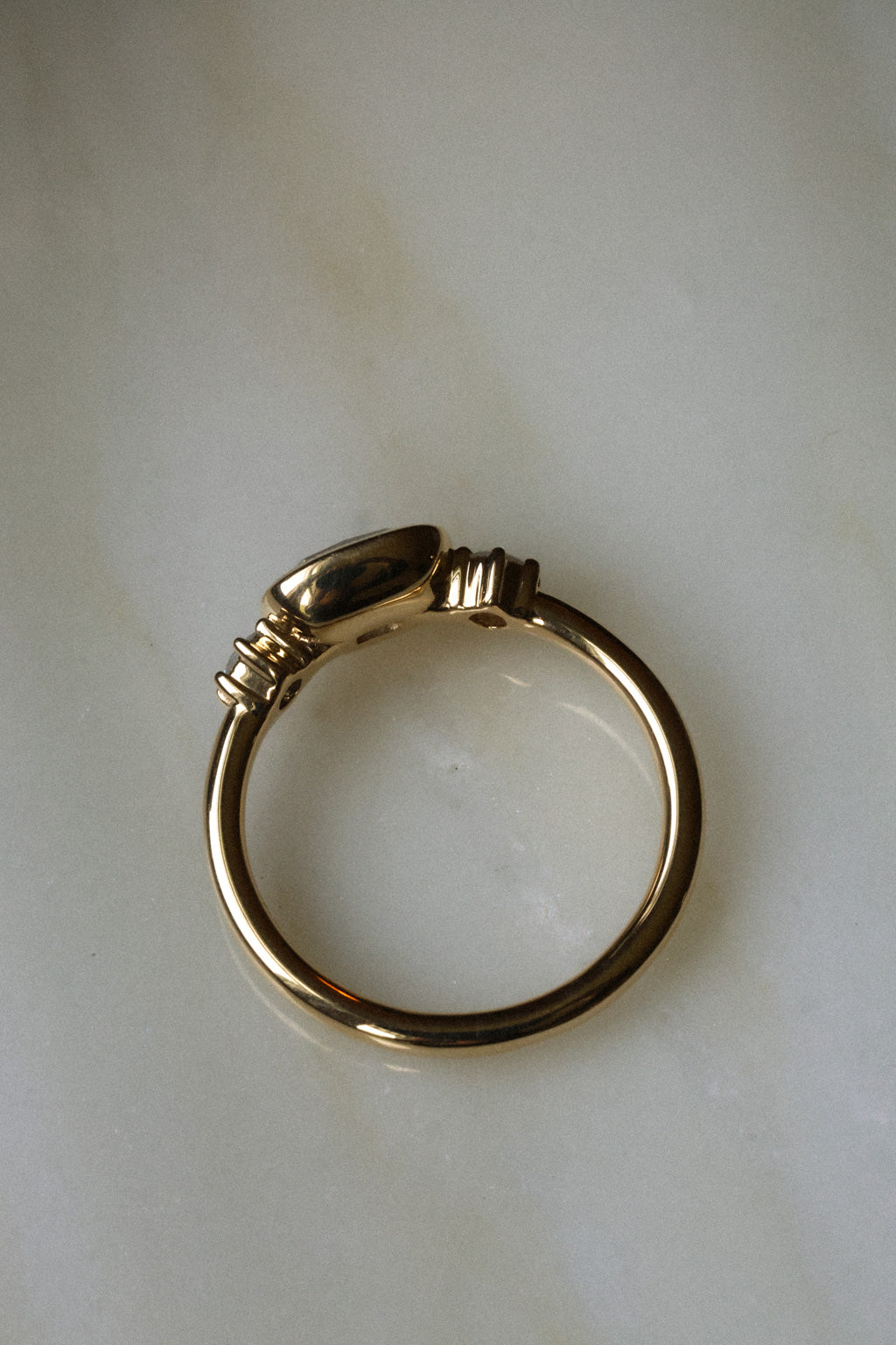 Photo of an engagement ring from above on a pale marble surface, showing the full circle of the band and the smooth polished gold back of the setting. The center stone is not visible from this angle. The ring is a thin, fully rounded yellow gold band with the bezel and side stone settings seen from behind.