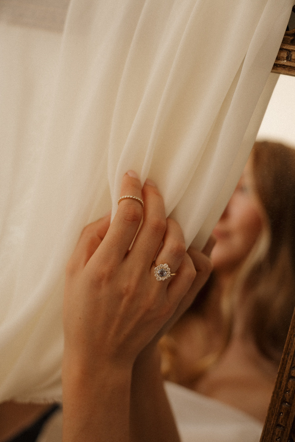A hand gathering a sheer chiffon curtain draped over a gilded mirror, wearing a pale blue sapphire halo engagement ring and a slim diamond band on the index finger, with a woman's reflection softly blurred in the mirror