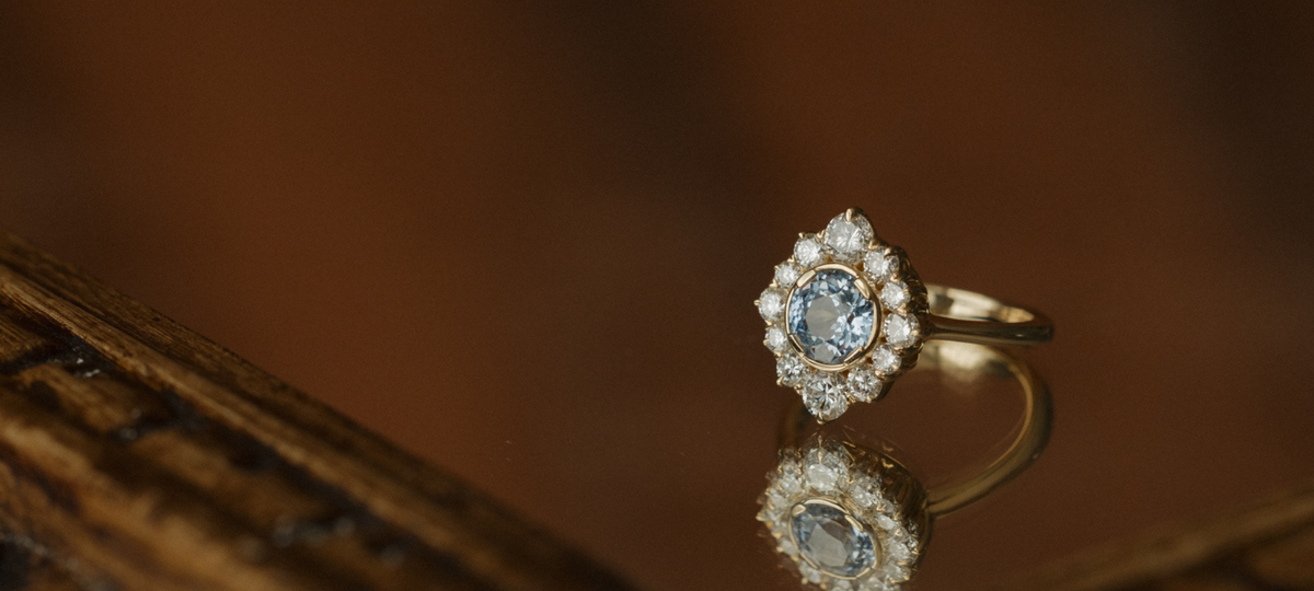 Angled view of a yellow gold engagement ring with a round blue Ceylon sapphire in a bezel setting, ringed by an antique-style halo of round diamonds, reflected on a dark glossy mirror surface beside the edge of the antique brass frame.
