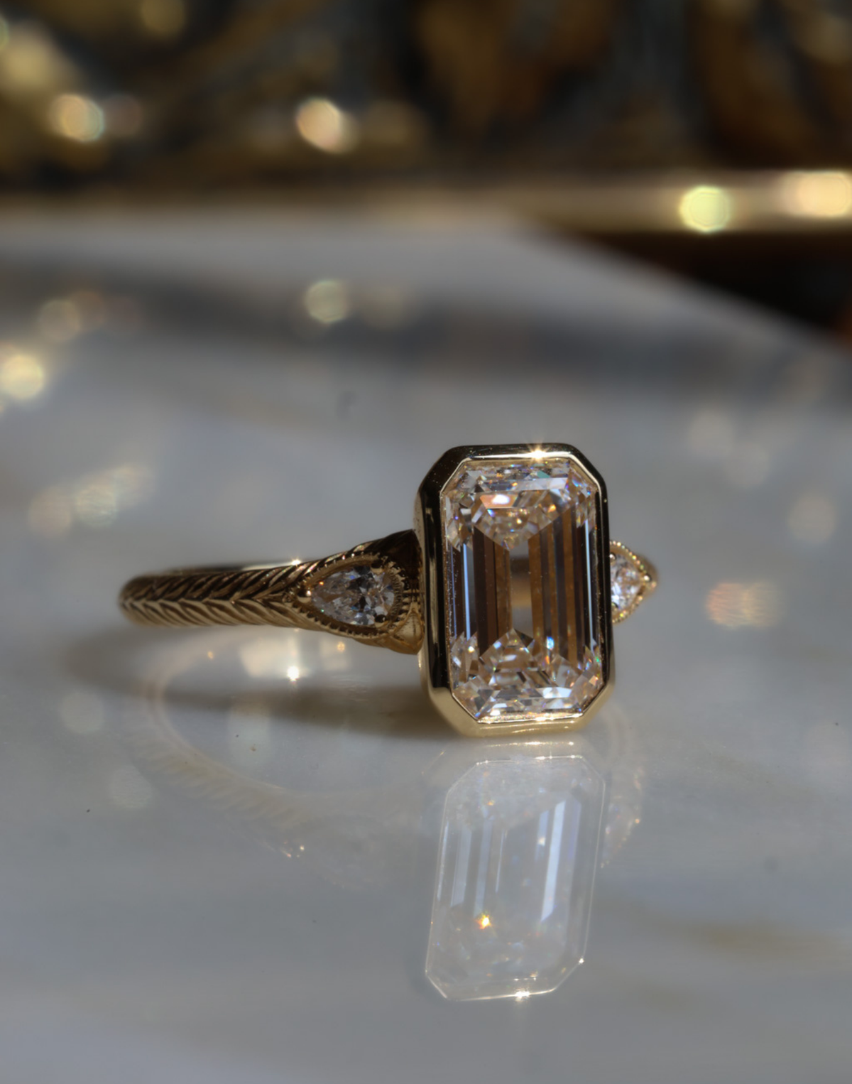 Angled front view of a yellow gold engagement ring with an emerald cut diamond in a bezel setting, a milgrain-edged pear cut diamond on each side and a hand-engraved wheat band, on a reflective cream marble surface with an antique-style brass jewelry box blurred behind.