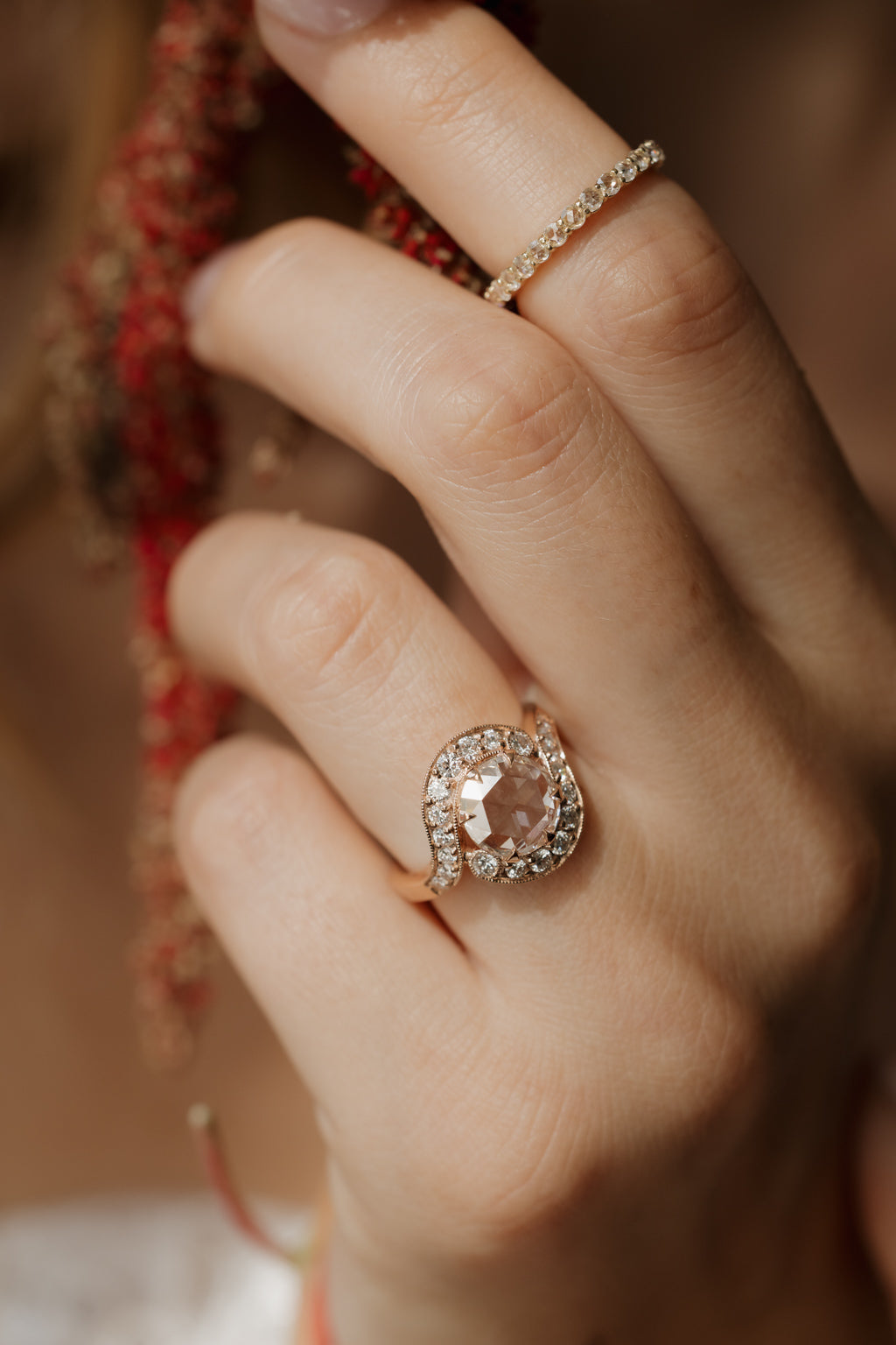 Close up photo of a hand wearing an engagement ring on the ring finger, turned so the setting faces the camera, with dried red amaranth out of focus behind. A thin diamond eternity band sits on the index finger. The ring is a rose gold curved bypass setting with a round rose cut diamond in a halo of single cut diamonds.