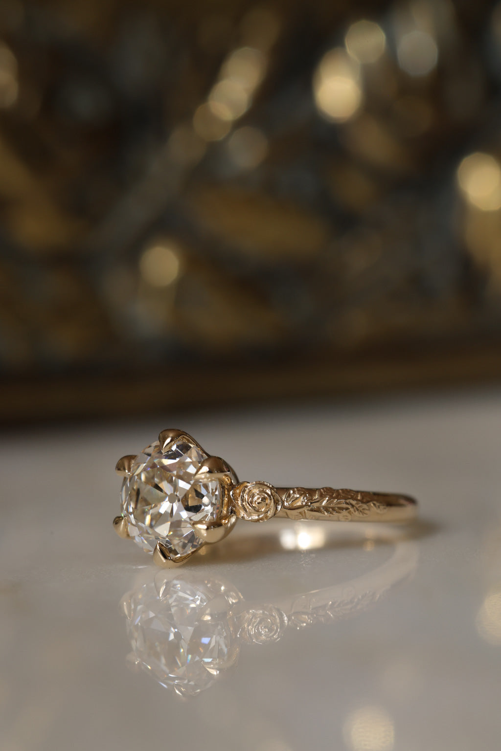 Angled side view of a yellow gold engagement ring with an old European cut diamond held by six claw prongs, a sculpted rose at the shoulder, and a vine motif along the band, on a reflective cream marble surface with an antique-style brass jewelry box blurred behind.