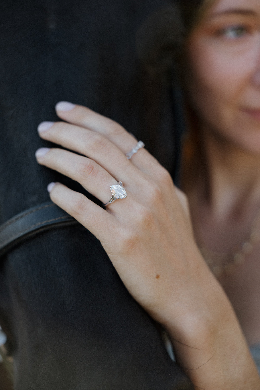 On-hand view of a white gold engagement ring with an old mine cut oval diamond and tapered baguette side stones, worn on the ring finger with a diamond band worn on the index finger, the hand resting on a black horse beside a leather bridle strap.