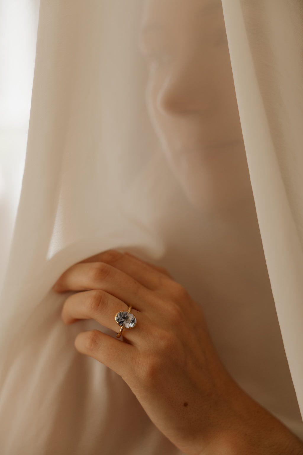 Photo of a hand pressed into a sheer cream curtain, a woman standing behind it, wearing an engagement ring on the ring finger. The ring is a thin yellow gold band with an oval blue Ceylon sapphire held in a collet setting.