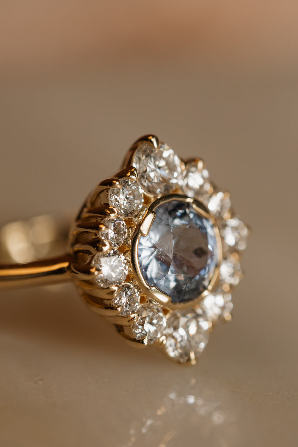 Close-up of a pale blue Ceylon sapphire set in a yellow gold bezel, ringed by round diamonds held in fluted claws, with soft reflections on the marble below.