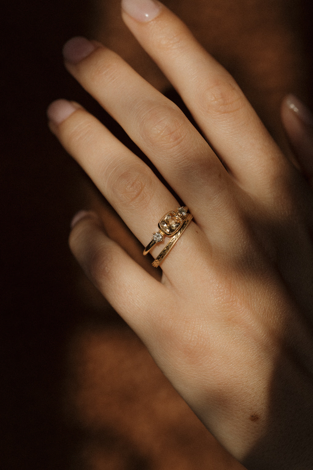 A hand in a streak of warm light against a dark background, wearing a yellow gold peach sapphire engagement ring stacked with a hand-engraved wedding band.