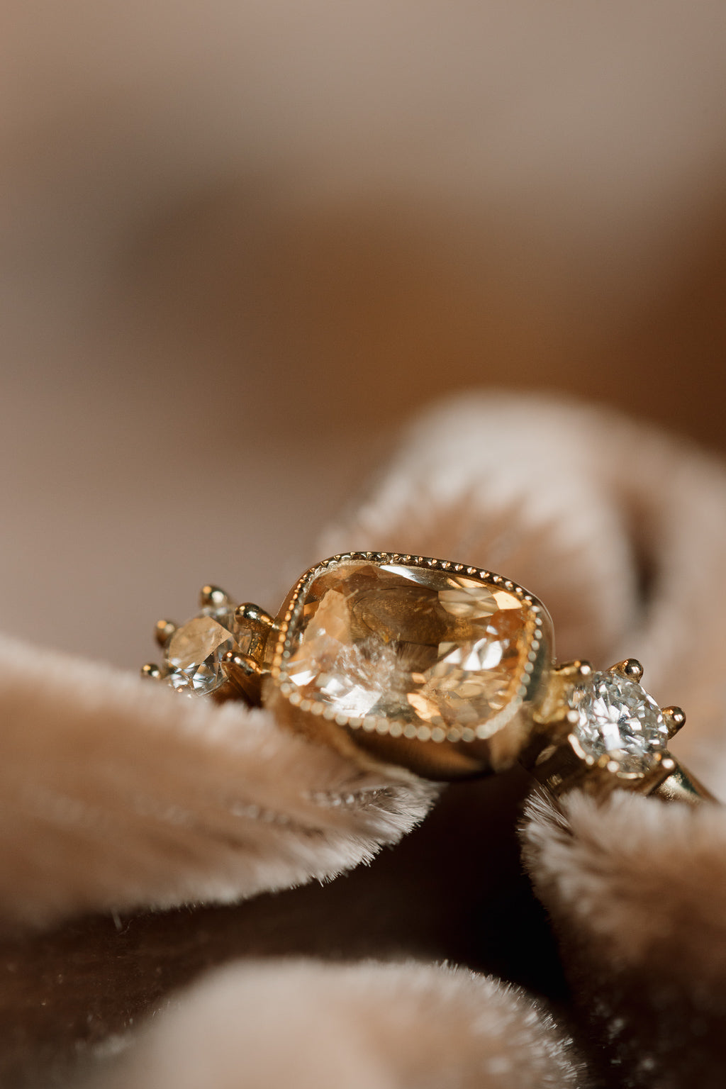Close-up of a yellow gold ring nestled in folds of blush velvet, showing a cushion cut peach sapphire in a beaded milgrain bezel and a round diamond in prongs on either side.