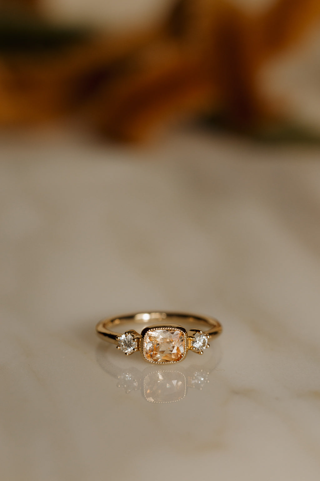 Front view of a yellow gold engagement ring with an east-west cushion cut peach sapphire in a milgrain bezel between two round diamonds, on a pale marble surface with its reflection below and warm blurred tones behind.