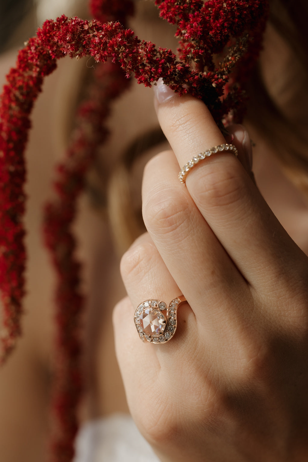 Photo of a hand reaching up to touch hanging stems of dried red amaranth, wearing an engagement ring on the ring finger and a thin diamond eternity band on the index finger. The ring is a rose gold curved bypass setting with a round rose cut diamond in a halo of single cut diamonds.