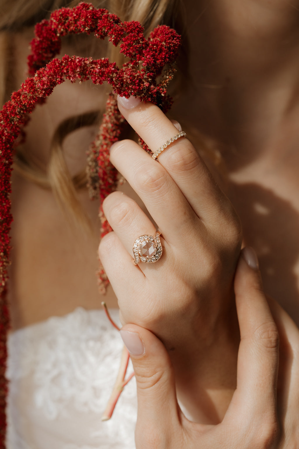 Photo of a woman in a white lace dress holding stems of dried red amaranth near her face, out of focus, with her hand in focus in the foreground. She wears an engagement ring on the ring finger and a thin diamond eternity band on the index finger. The ring is a rose gold curved bypass setting with a round rose cut diamond in a single cut diamond halo.