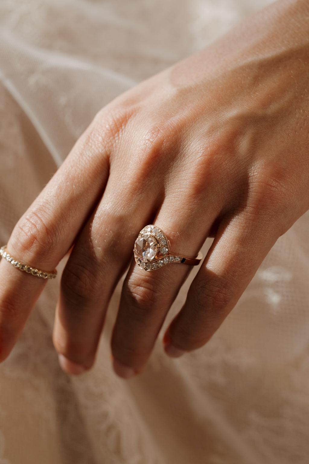Photo of a hand resting on sheer pale fabric at a side angle in soft shadow, wearing an engagement ring on the ring finger, with a diamond eternity band on the other hand at the edge of the frame. The ring is a rose gold curved bypass setting with a round rose cut diamond in a halo of single cut diamonds.