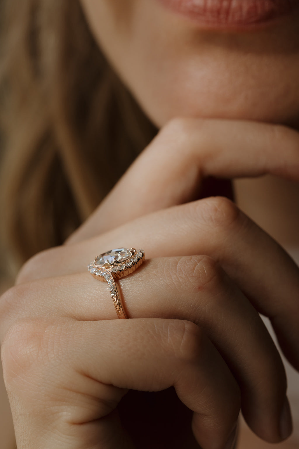 A close up of a woman with her hand under her chin, she is wearing a rose gold engagement ring on her hand. The engagement ring has a double prong set oval rose cut diamond with a single cut bypass halo surrounding it.