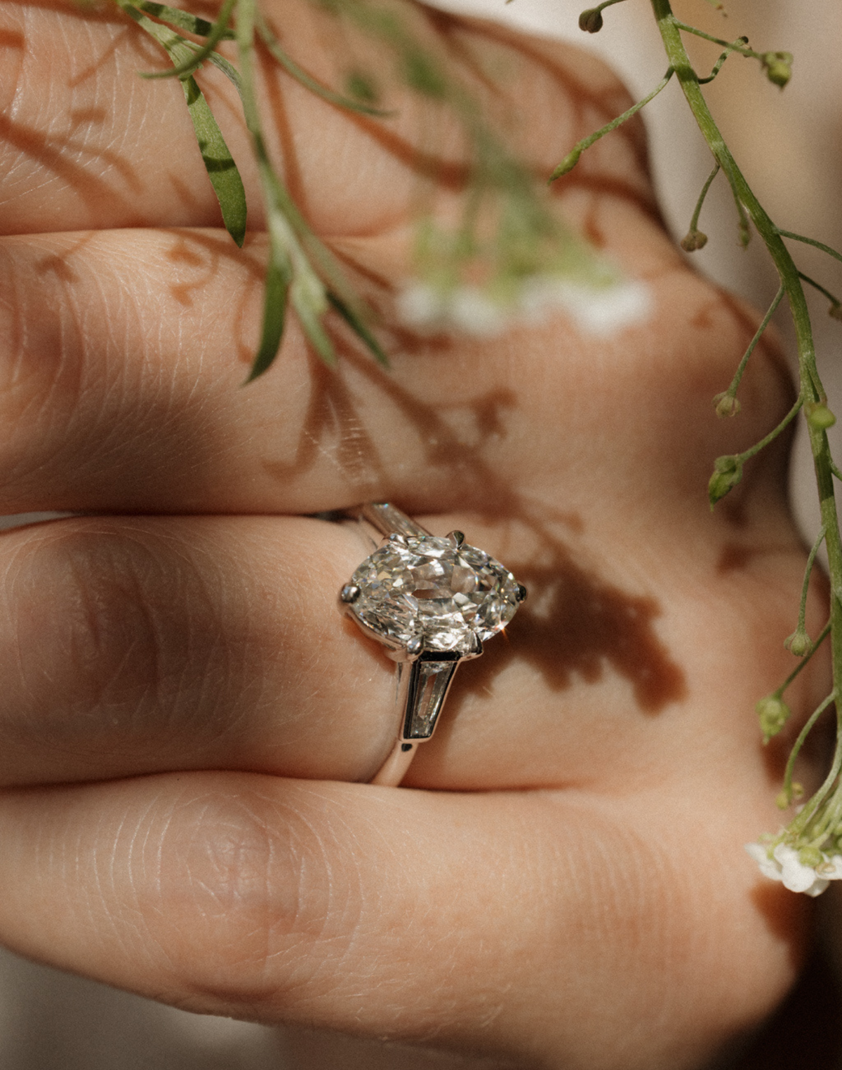 Close view of a hand wearing a white gold engagement ring with an elongated old mine oval diamond in claw prongs and a tapered baguette diamond on each side, with sprigs of small white wildflowers and their shadows falling across the fingers.