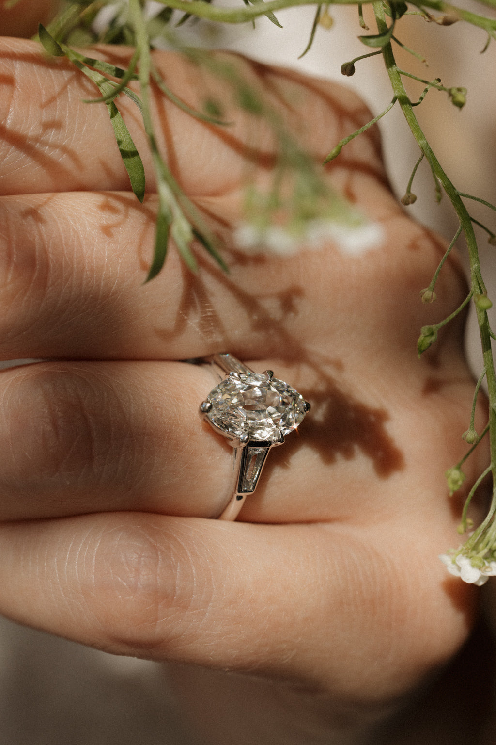 Close-up on hand of a white gold engagement ring with an old mine cut oval diamond in six claw prongs and a tapered baguette side stone, with small white wildflowers casting shadows across the fingers.