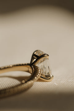 Close side profile of a yellow gold engagement ring showing an antique emerald cut diamond held by curved claw prongs above a ribbed gallery and a milgrain-edged hand-engraved band, on cream woven linen in warm light.