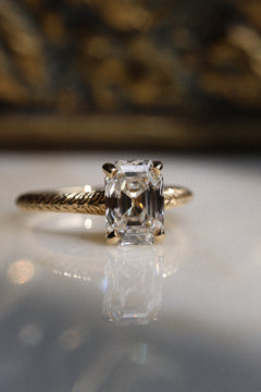 Front view of a yellow gold engagement ring with an antique emerald cut diamond in four claw prongs on a hand-engraved band, on a reflective cream marble surface with an antique-style brass jewelry box blurred behind.