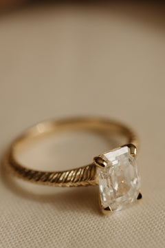Angled view of a yellow gold engagement ring with an antique emerald cut diamond in four claw prongs on a hand-engraved band, on cream woven linen.