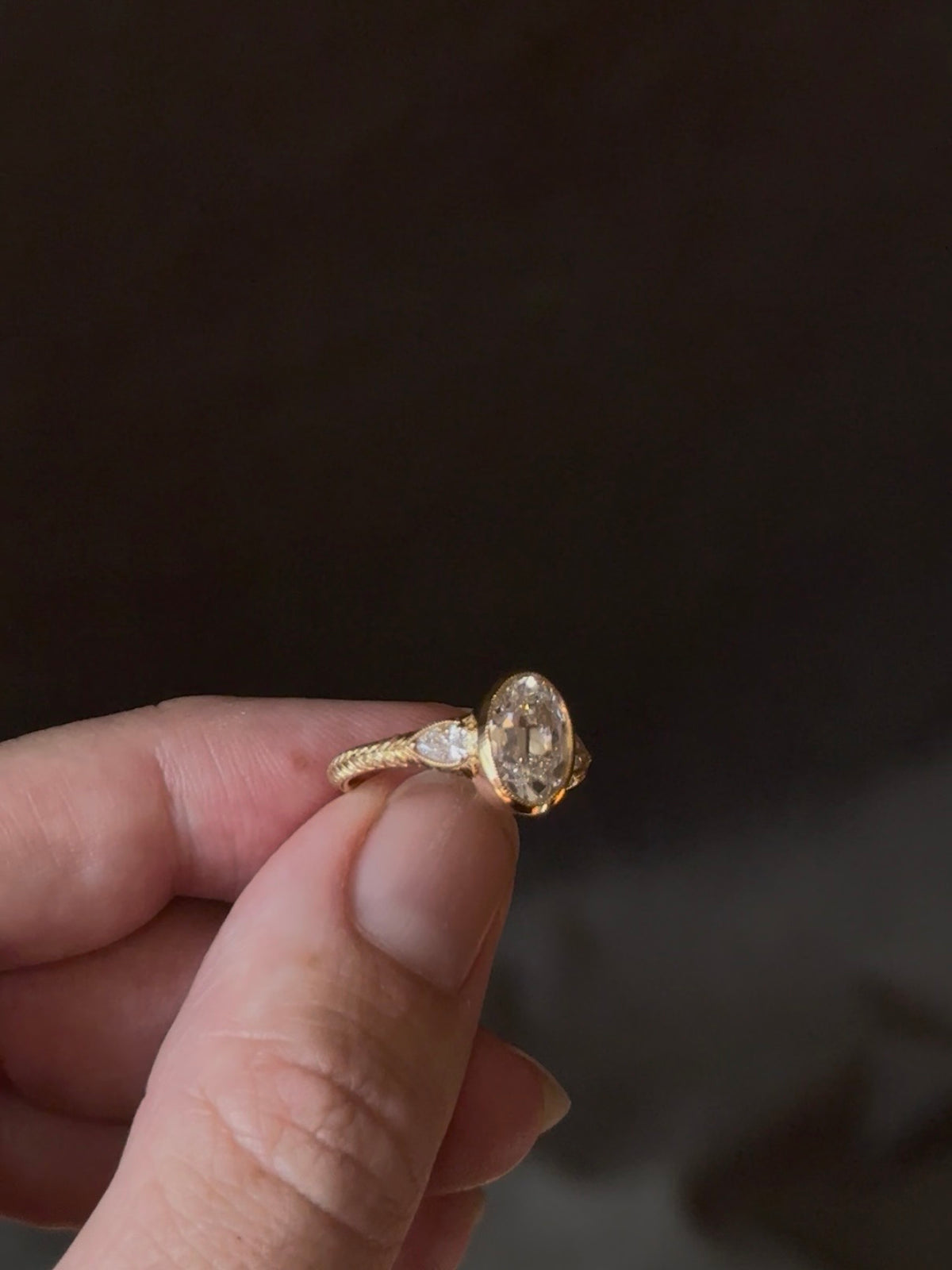 Close view of fingers holding a yellow gold engagement ring with a bezel-set antique oval diamond, pear cut diamonds at the shoulders and a wheat hand-engraved band, against a dark background.