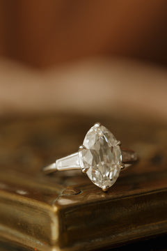 Front view of a white gold engagement ring with an old mine cut oval diamond in six claw prongs and bezel-set tapered baguette side stones, resting on the corner of an antique-style brass jewelry box in warm light.