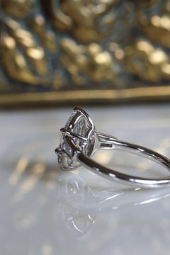 Side view of a white gold engagement ring with the old mine cut oval diamond facing left, showing the six-prong setting and a tapered baguette side stone along the slim polished band, on a white reflective surface before a blurred gilded jewelry box.