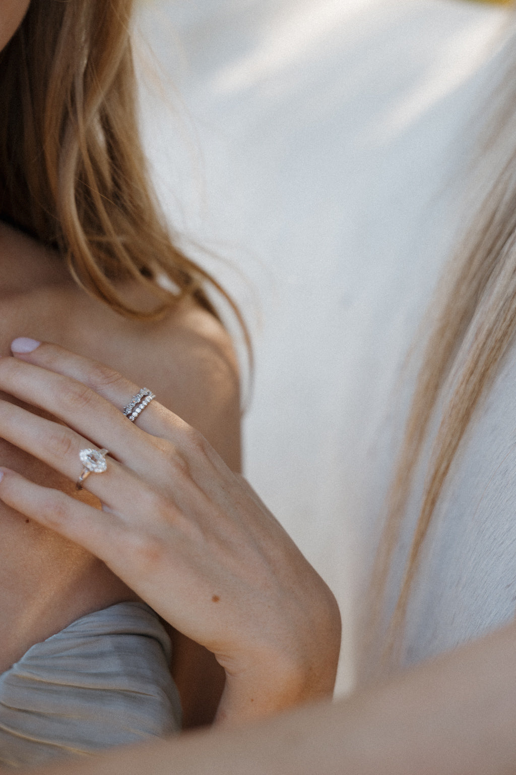 On-hand view of a white gold engagement ring with an old mine cut oval diamond, worn with two stacked diamond bands on the middle finger, the hand resting at the collarbone beside a pale blue dress with a white horse in the background.