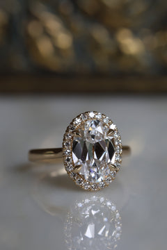 Front view of a yellow gold engagement ring with an antique cut oval diamond in four soft V-prongs inside a single cut diamond halo, on a reflective cream marble surface with an antique-style brass jewelry box blurred behind.
