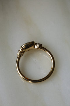 Photo of an engagement ring from above on a pale marble surface, showing the full circle of the band and the smooth polished gold back of the setting. The center stone is not visible from this angle. The ring is a thin, fully rounded yellow gold band with the bezel and side stone settings seen from behind.