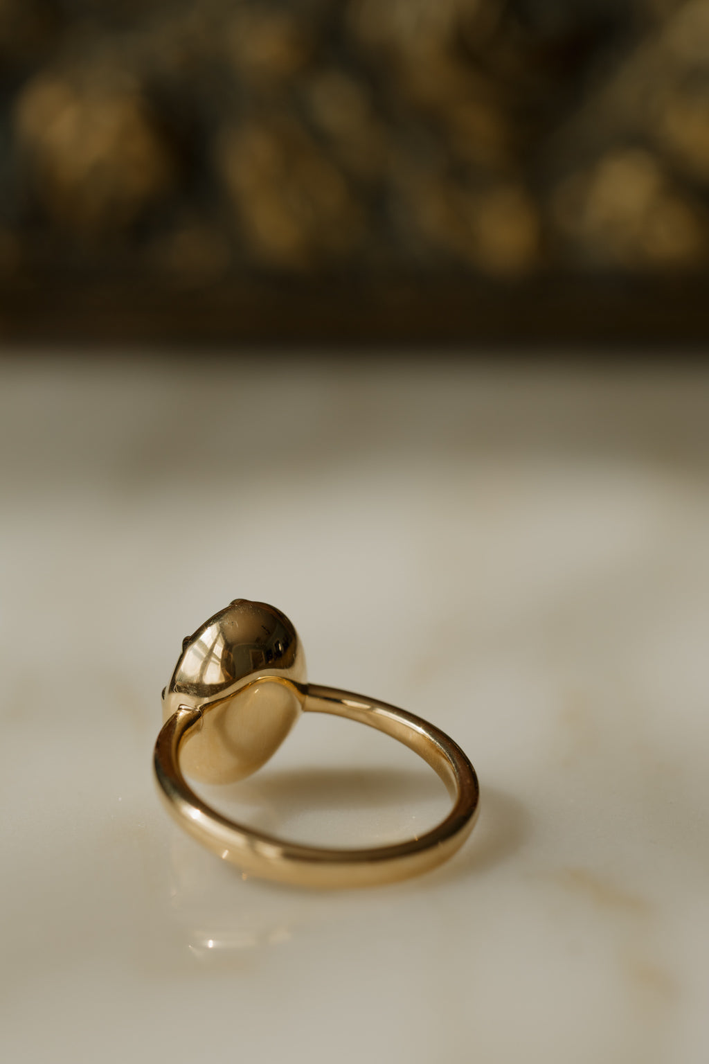 Close up photo of the back of an engagement ring on a white surface, showing the smooth gold underside of the setting. The ring is a thin yellow gold band, and the collet cup holding the oval sapphire is closed and polished underneath.