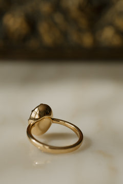 Close up photo of the back of an engagement ring on a white surface, showing the smooth gold underside of the setting. The ring is a thin yellow gold band, and the collet cup holding the oval sapphire is closed and polished underneath.