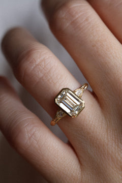Close up of a hand wearing a bezel set emerald cut diamond engagement ring. The ring has pear cut accent stones with a milgrain detail. The band is yellow gold and hand engraved band. 