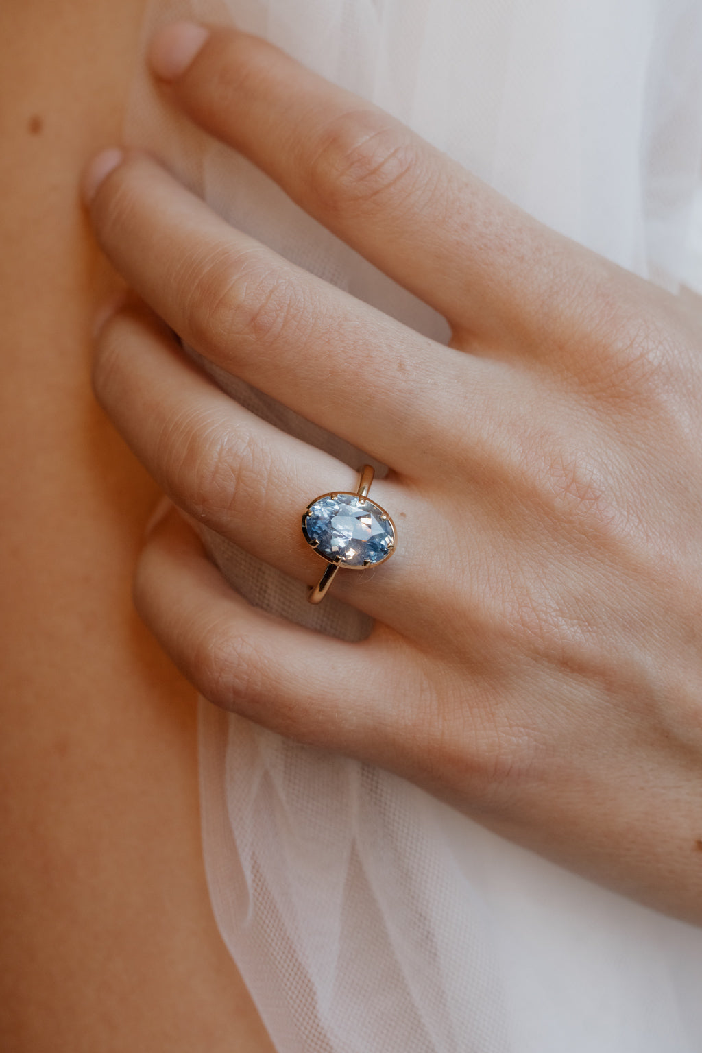 Close up photo of a hand resting against a bare shoulder over white fabric, wearing an engagement ring on the ring finger. The ring is a thin yellow gold band with an oval blue Ceylon sapphire held in a collet setting.