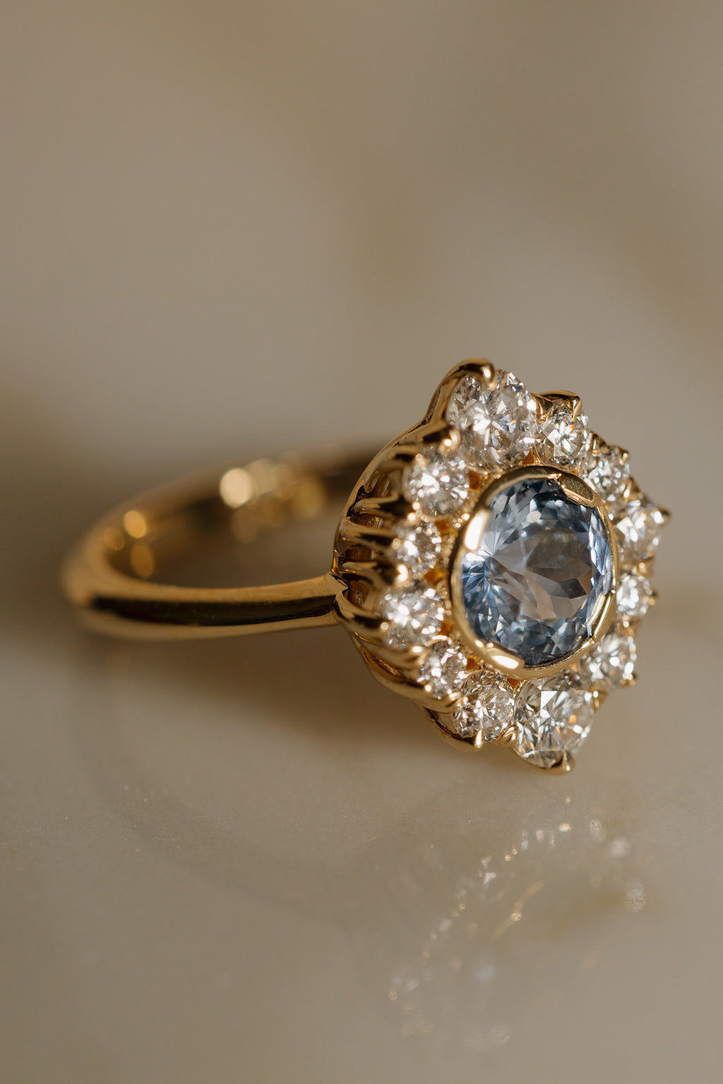 Angled view of a yellow gold engagement ring with a round pale blue Ceylon sapphire in a bezel, surrounded by an elongated antique-style halo of round diamonds with fluted gold claws along the side, on a pale marble surface.