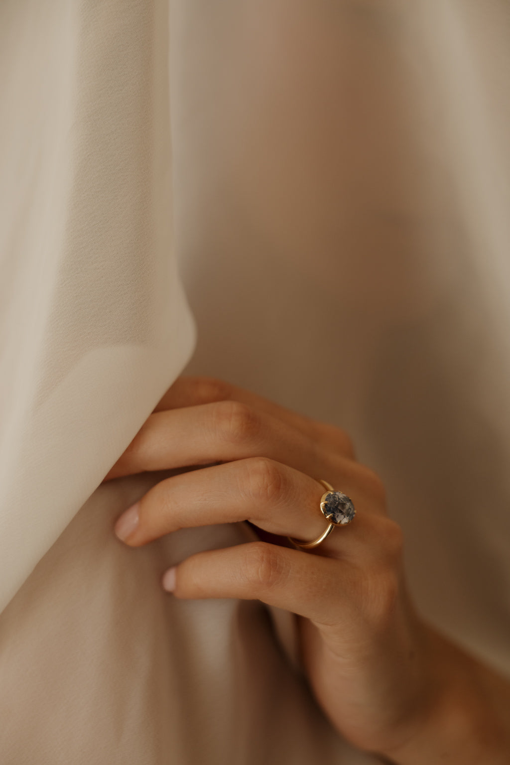 Photo of a hand resting against a draped sheer cream curtain, wearing an engagement ring on the ring finger. The ring is a thin yellow gold band with an oval blue Ceylon sapphire in a collet setting. Most of the frame is soft folds of pale fabric.
