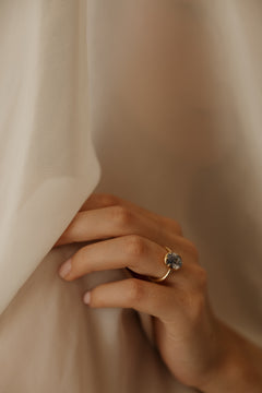 Photo of a hand resting against a draped sheer cream curtain, wearing an engagement ring on the ring finger. The ring is a thin yellow gold band with an oval blue Ceylon sapphire in a collet setting. Most of the frame is soft folds of pale fabric.