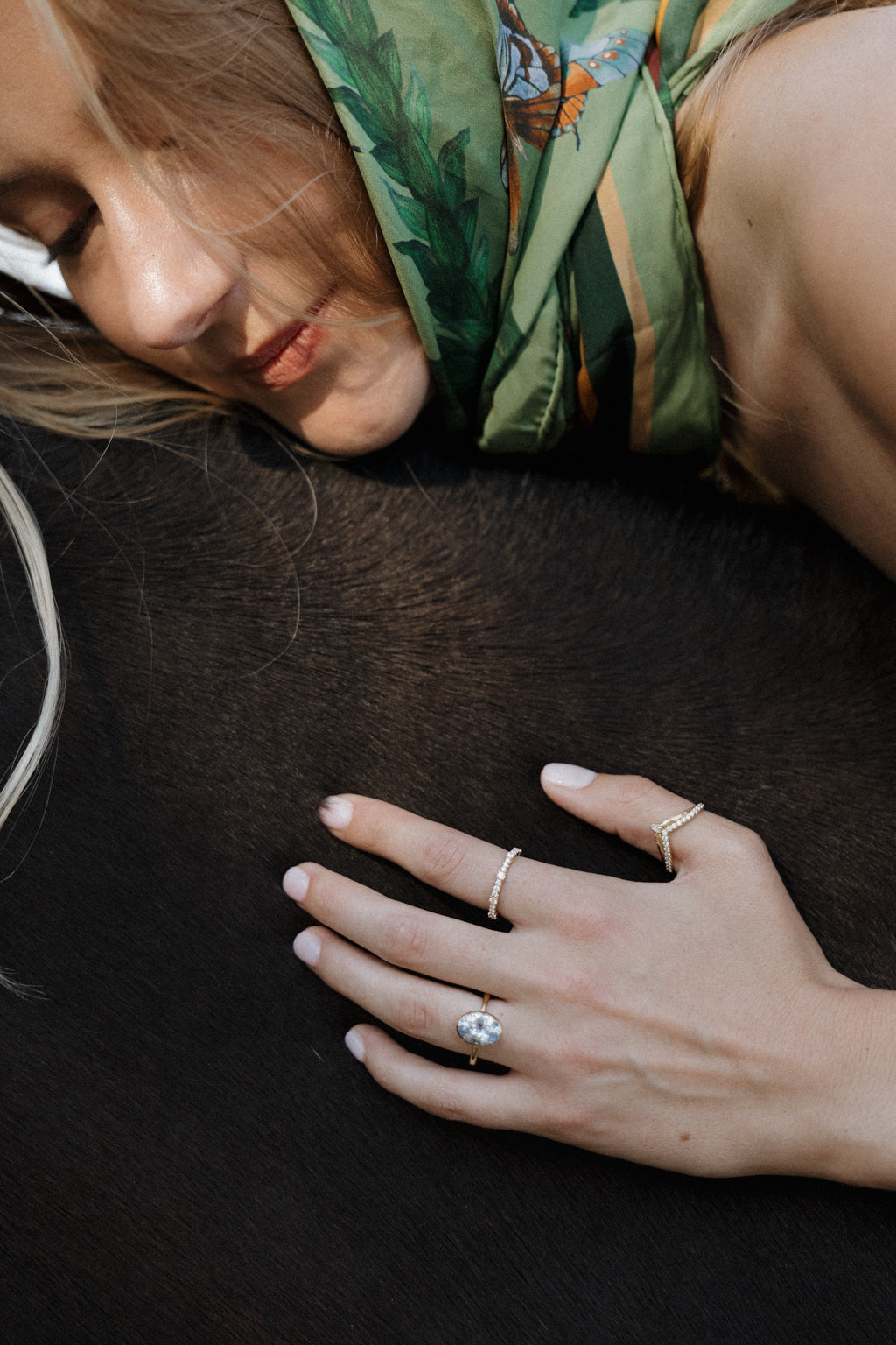 Photo of a woman lying face down across the back of a dark brown horse, her blonde hair and a green printed silk scarf in the frame, with both hands resting on the horse. She wears an engagement ring with an oval blue Ceylon sapphire in a yellow gold collet setting, and thin diamond bands on two other fingers.