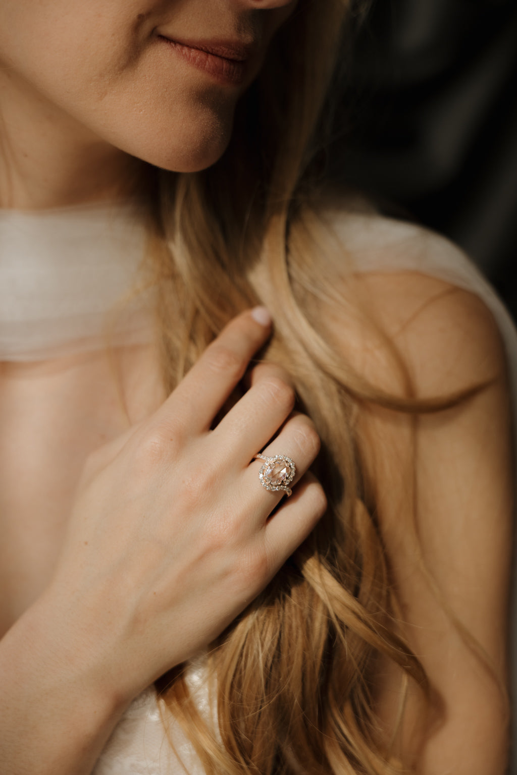 A woman partially in frame is wearing a wedding dress and is reaching towards her white tulle scarf around her neck. Her hand has a rose gold engagement ring, the ring has an oval shaped rose cut diamond center stone with a bypass halo made up of twenty-eight single cut diamonds. The band has engraved details where the halo ends.