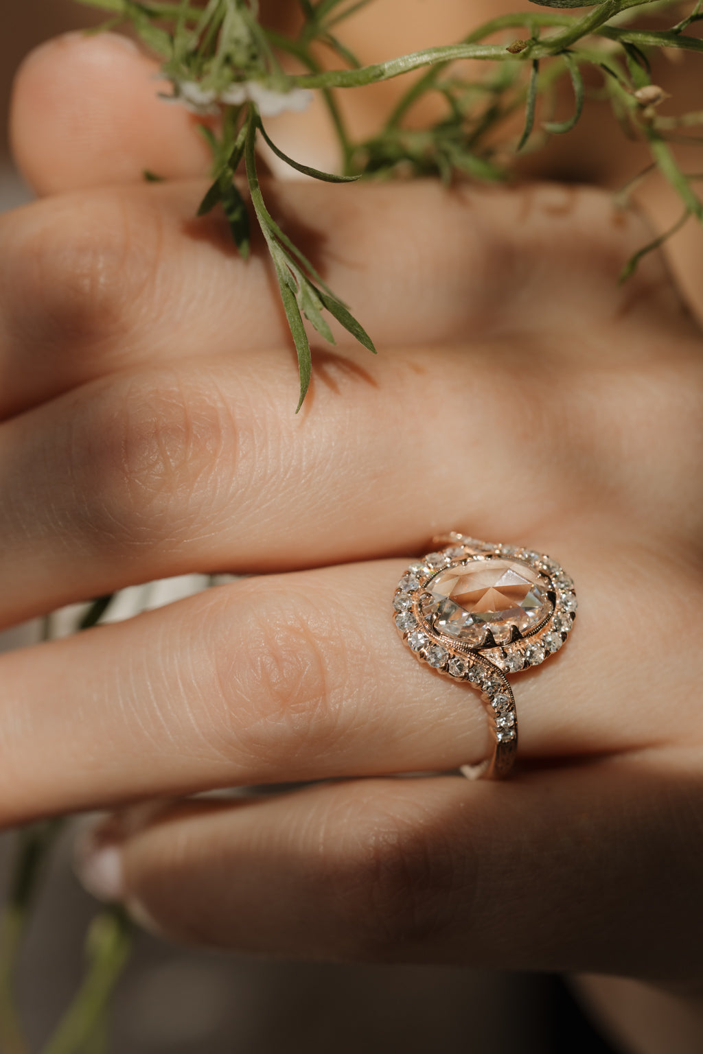 Side view of a hand holding a green plant, the hand is wearing an oval rose cut diamond engagement ring. The ring is rose gold with engravings on the band, there is a single cut diamond bypass halo. 