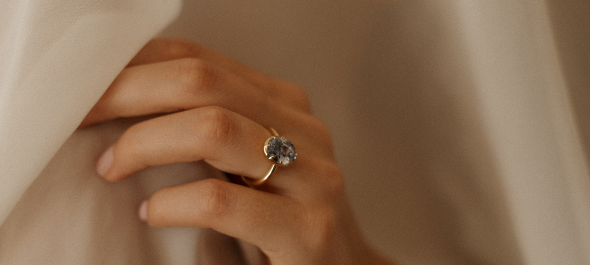 Close view of a hand resting against cream silk, wearing a yellow gold engagement ring with an oval pale blue Ceylon sapphire held in an eight-prong collet setting on a slim band.