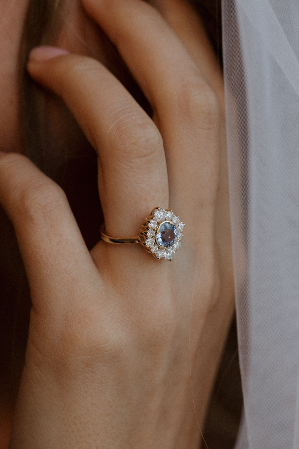 Close view of a hand raised near the face, wearing a yellow gold engagement ring with a pale blue Ceylon sapphire in an antique-style diamond halo, with a sheer tulle veil falling alongside.