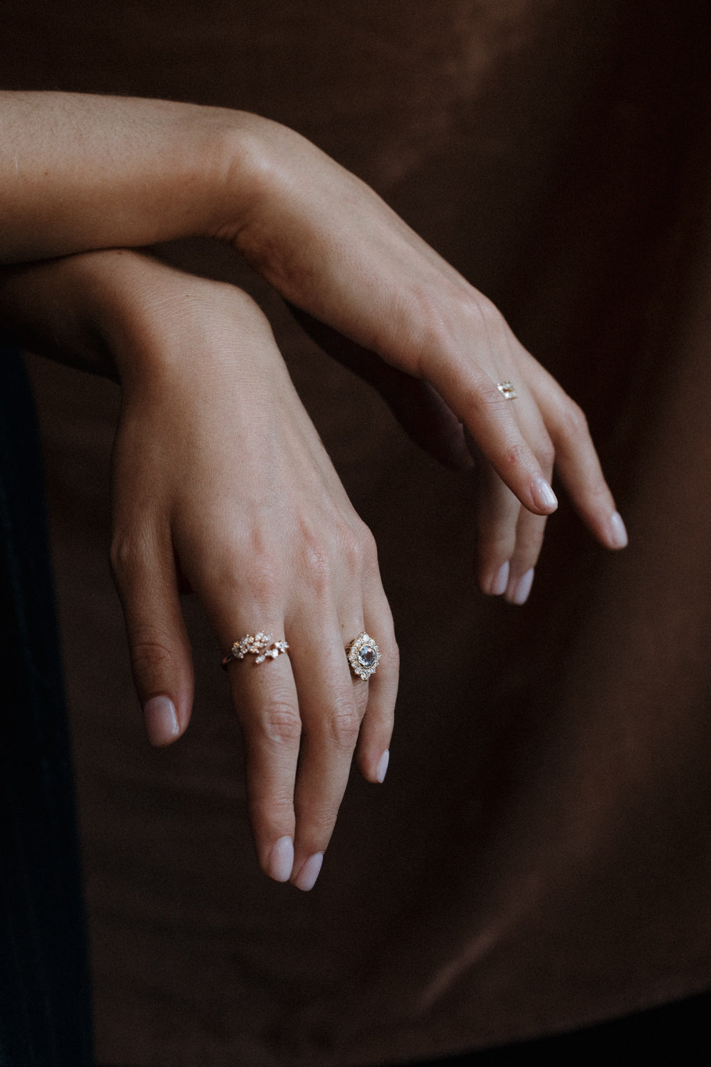 Two crossed hands resting one over the other against a dark brown backdrop, as if leaning on something out of frame. One hand wears a yellow gold engagement ring with a pale blue Ceylon sapphire in an antique-style diamond halo and a diamond cluster band on the index finger; the other wears a small diamond ring.