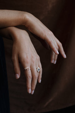 Two crossed hands resting one over the other against a dark brown backdrop, as if leaning on something out of frame. One hand wears a yellow gold engagement ring with a pale blue Ceylon sapphire in an antique-style diamond halo and a diamond cluster band on the index finger; the other wears a small diamond ring.