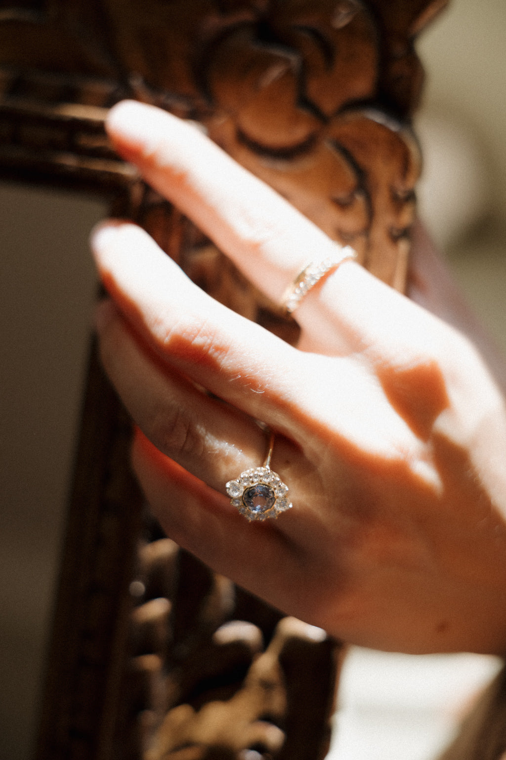 A hand resting against a carved gilded mirror frame in a streak of sunlight, wearing a yellow gold engagement ring with a pale blue sapphire in a diamond halo and a diamond band on the middle finger.