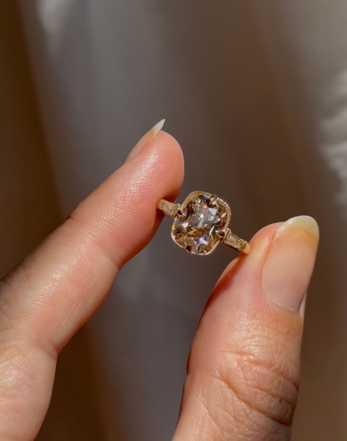 Close view of fingertips holding a yellow gold engagement ring with a champagne old mine cut diamond in four claw prongs, bordered in hand-dotted milgrain, with a tapered baguette diamond on each side, against a soft blurred brown background.