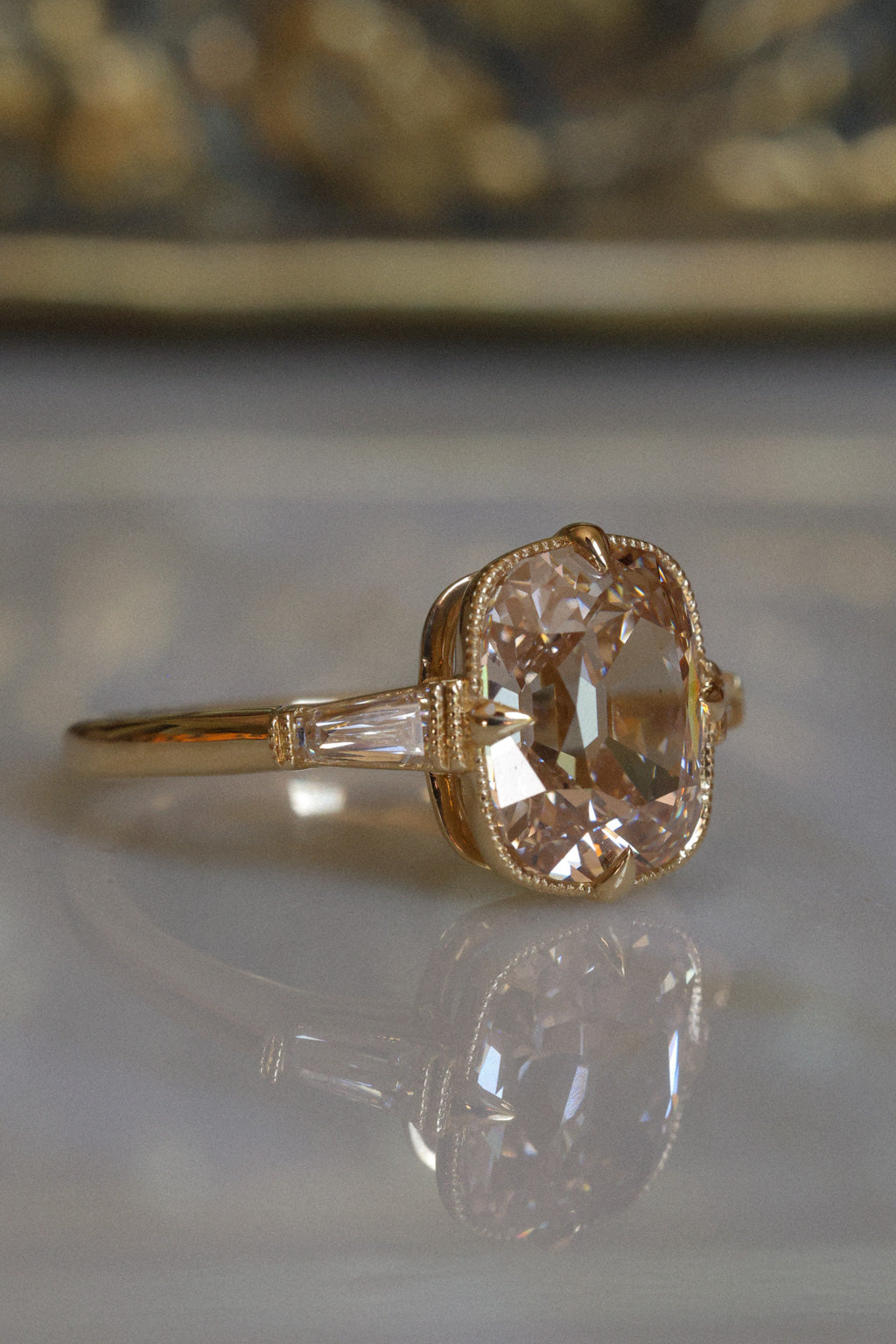 Close side view of a yellow gold engagement ring with a champagne old mine cut diamond held in claw prongs and edged in milgrain, with a tapered baguette diamond on each side, resting on a reflective cream marble surface with an antique-style brass jewelry box blurred behind it.