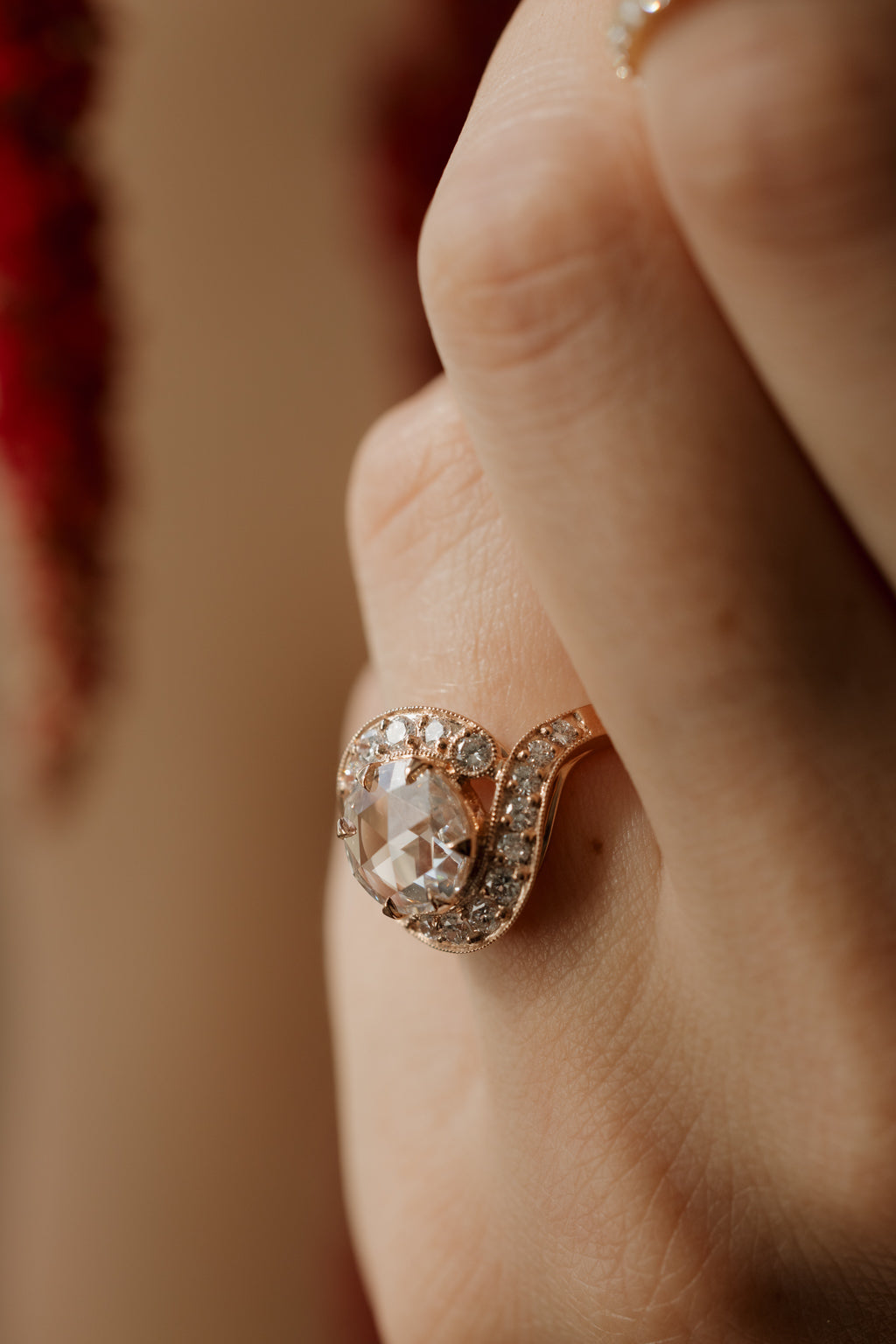 Very close up photo of an engagement ring worn on an upright hand, with dried red amaranth out of focus at the edge of the frame. The ring is a rose gold curved bypass band. It has a round rose cut diamond held in prongs, ringed by a halo of single cut diamonds with milgrain along the edges.
