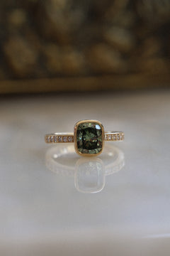 Front view of a yellow gold engagement ring with a cushion cut green diamond in a slim milgrain bezel on a diamond-set band, on a reflective cream marble surface with an antique-style brass jewelry box blurred behind.