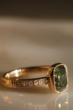 Angled side view of a yellow gold engagement ring with a cushion cut green diamond in a slim milgrain bezel, a row of diamonds set along the band between milgrain edges, and a grooved band detail, on a reflective cream marble surface in warm light.